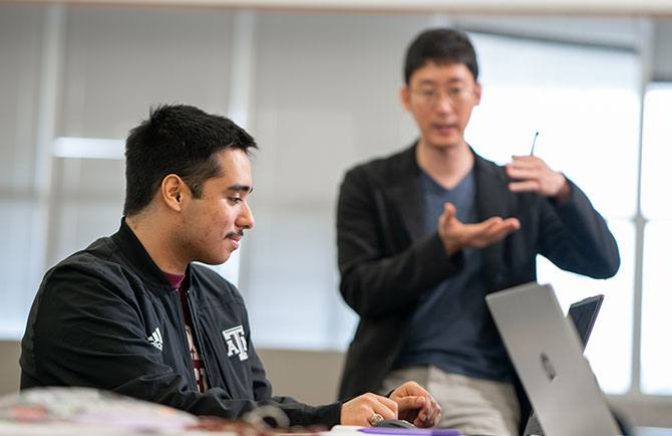 A professor instructs a student working at his laptop in a classroom